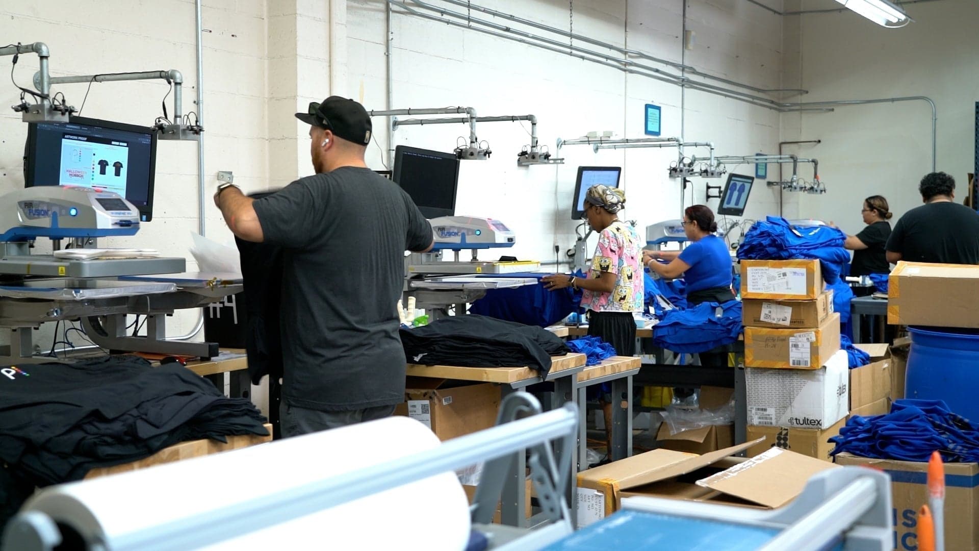 Workers operating DTF printers and handling blue garments in a commercial print shop facility